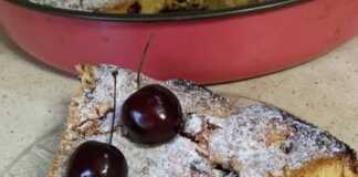 Μια φρεσκοψημένη, χρυσαφένια κερασόπιτα με πολλά κόκκινα κεράσια στην επιφάνεια, πασπαλισμένη με ζάχαρη άχνη, κομμένη σε κομμάτια.