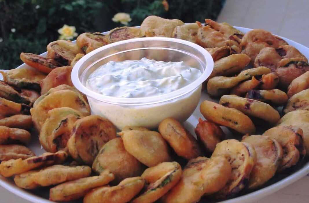 Ένα πιάτο με χρυσαφένιες τηγανιτές φέτες κολοκυθιού και μελιτζάνας δίπλα σε ένα μπολάκι με λευκό τζατζίκι.