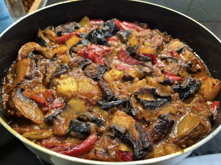 Ένα ταψί με λαχταριστά ψητά λαχανικά, όπως μελιτζάνες, πιπεριές και πατάτες, μέσα σε κόκκινη σάλτσα, με φρέσκα βότανα από πάνω.