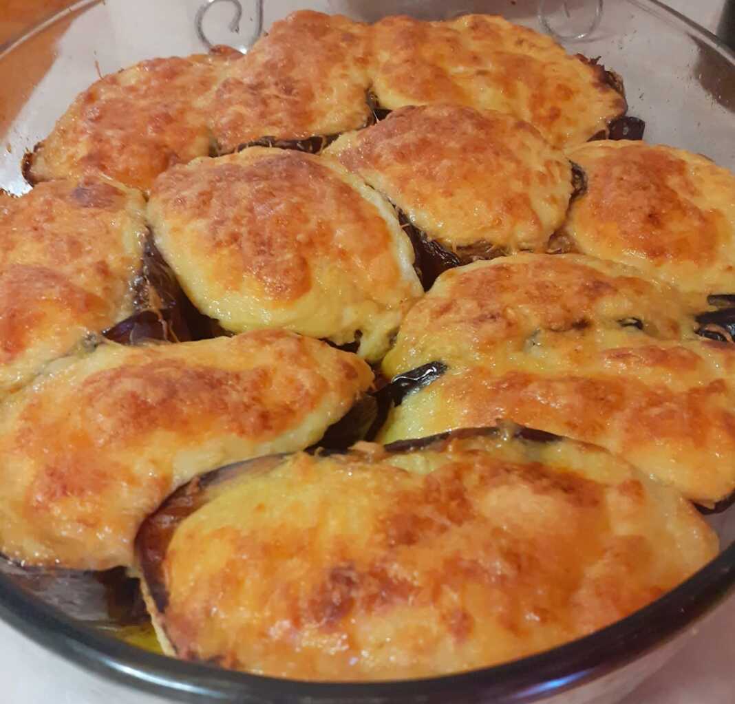 Ένα ταψί με γεμιστές μελιτζάνες σε σχήμα βαρκούλας, καλυμμένες με λευκή μπεσαμέλ και λίγο τριμμένο τυρί, ψημένες μέχρι να ροδίσουν.