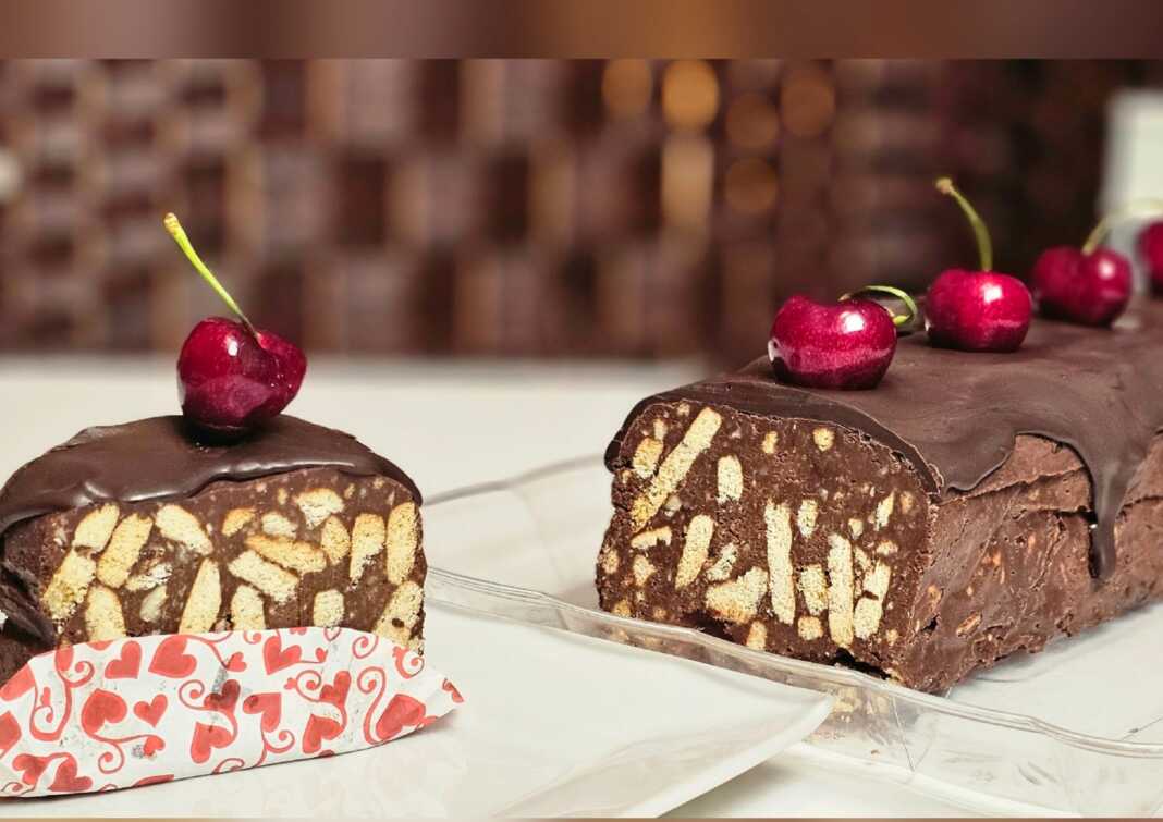 Ένα κομμένο κομμάτι από καλοκαιρινό μωσαϊκό με εμφανή κομμάτια μπισκότου, κερασιών και ξηρών καρπών, καλυμμένο με σοκολάτα, σερβιρισμένο σε λευκό πιάτο.