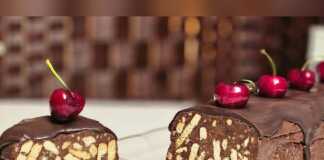 Ένα κομμένο κομμάτι από καλοκαιρινό μωσαϊκό με εμφανή κομμάτια μπισκότου, κερασιών και ξηρών καρπών, καλυμμένο με σοκολάτα, σερβιρισμένο σε λευκό πιάτο.