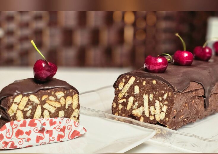 Ένα κομμένο κομμάτι από καλοκαιρινό μωσαϊκό με εμφανή κομμάτια μπισκότου, κερασιών και ξηρών καρπών, καλυμμένο με σοκολάτα, σερβιρισμένο σε λευκό πιάτο.