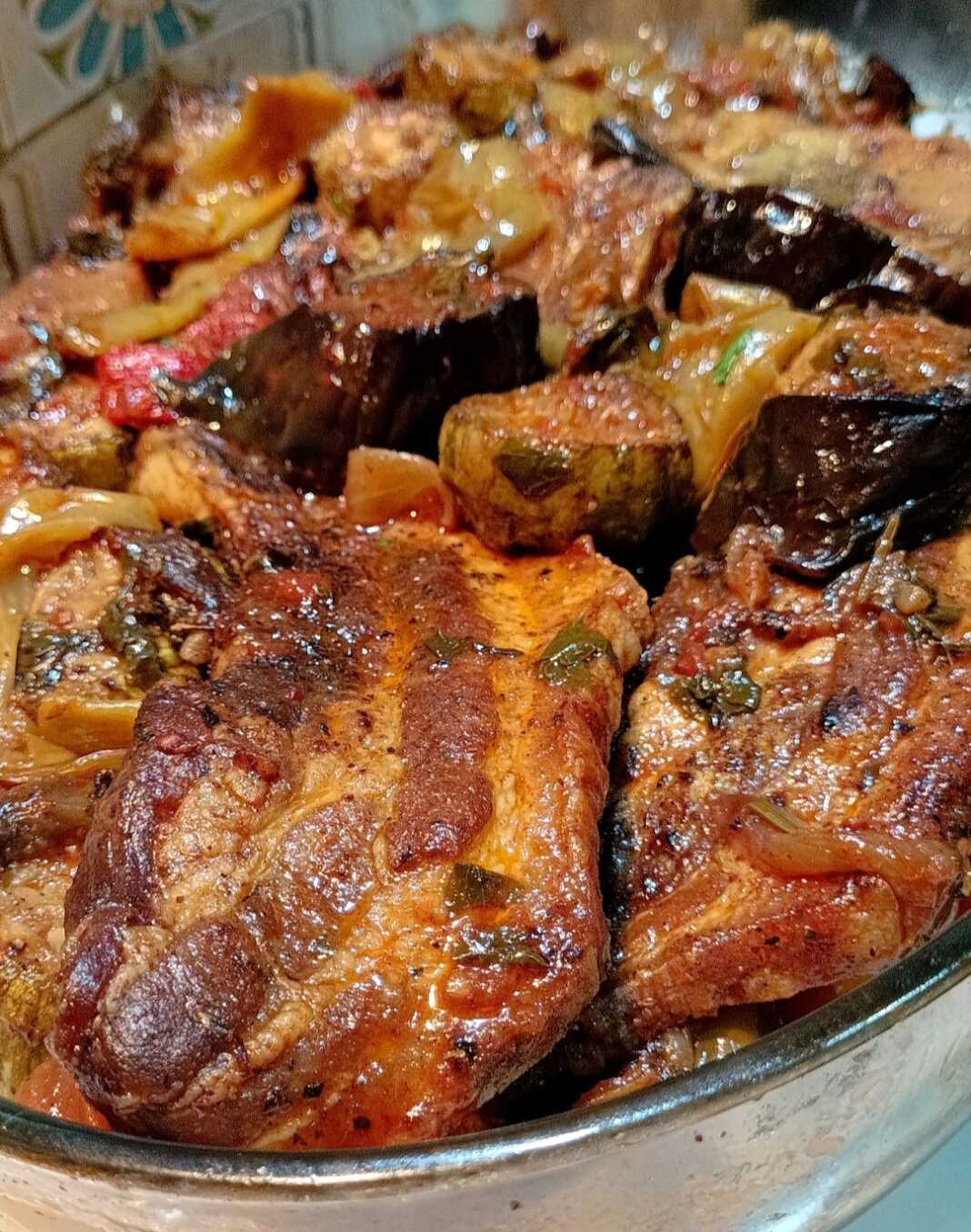 Ένα ταψί γεμάτο με πολύχρωμα ψητά λαχανικά, όπως κολοκυθάκια, μελιτζάνες, πατάτες και πιπεριές, αναμεμειγμένα με ζουμερές ροδισμένες πανσέτες χοιρινό.