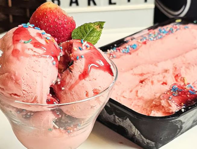 pagoto fraoula1 Ένα μπολ γεμάτο αφράτο, ροζ παγωτό φράουλα, διακοσμημένο με φρέσκες φράουλες και σιρόπι, έτοιμο για σερβίρισμα.