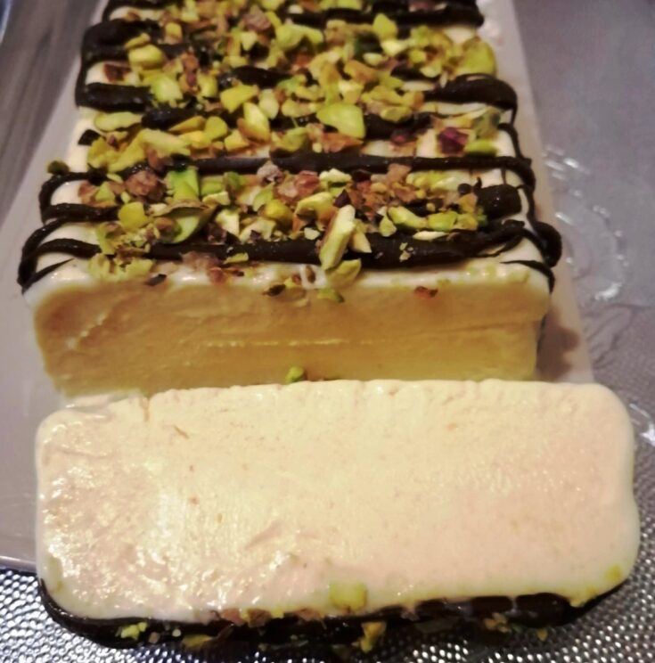 Μια φέτα από παγωτό Σεμιφρέντο με κρεμώδη υφή, γαρνιρισμένη με λιωμένη σοκολάτα και ψιλοκομμένα φιστίκια.