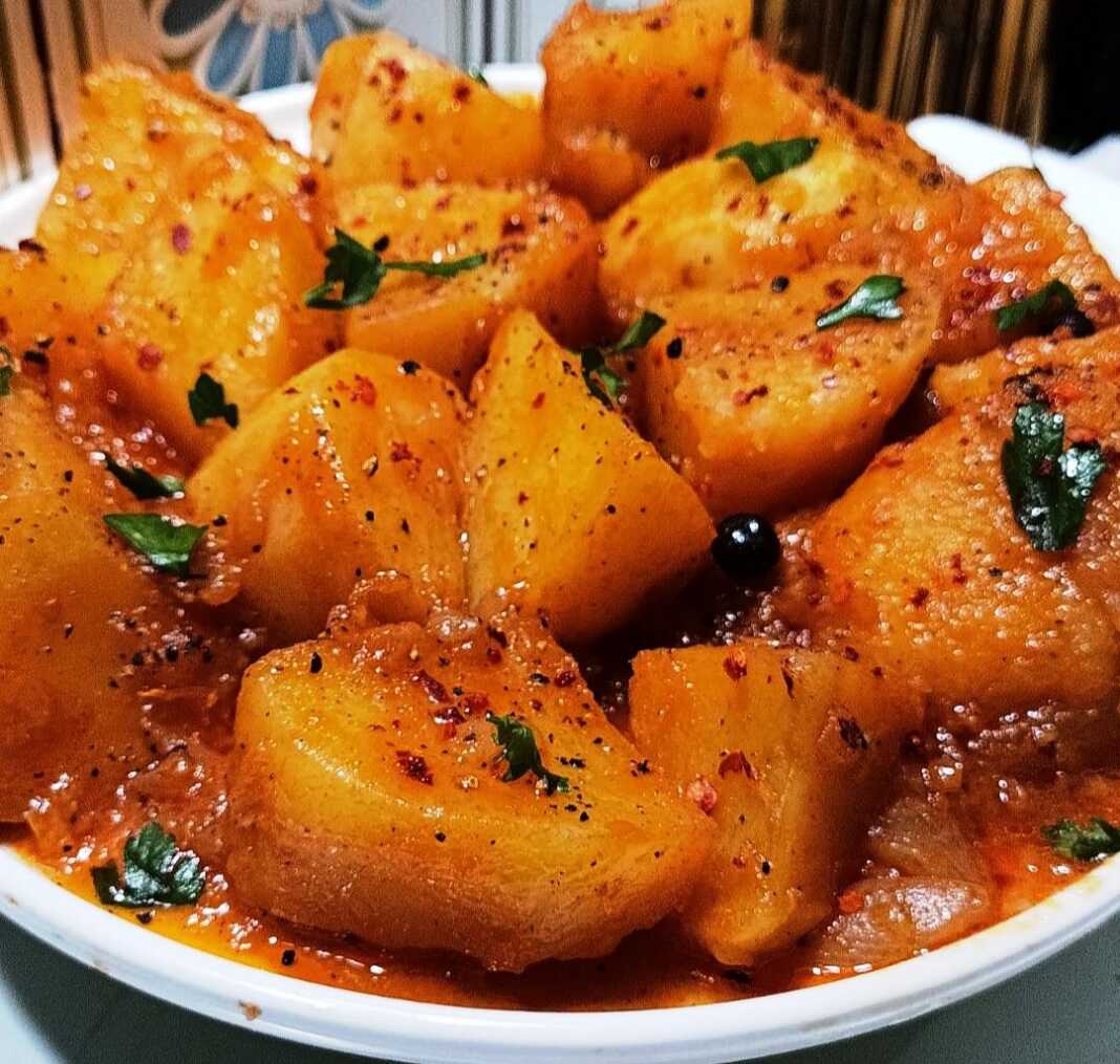 Ένα πιάτο με ζεστές πατάτες γιαχνί, περιχυμένες με κόκκινη σάλτσα, πασπαλισμένες με μαϊντανό και μπούκοβο, με ένα κομμάτι φέτας στο πλάι.