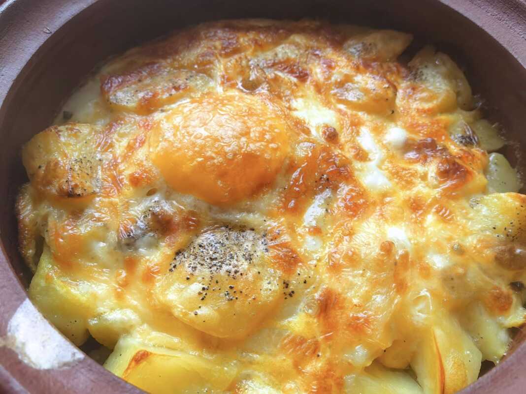 Ένα ταψί με χρυσαφένιες ψητές πατάτες, ανακατεμένες με τυρί, και δύο ολόκληρα αυγά στην επιφάνεια, ψημένα μέχρι να ροδίσουν.