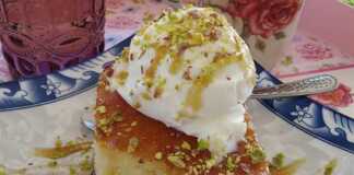 Ένα κομμάτι ραβανί, σιροπιασμένο, με χρυσαφένια επιφάνεια, σε ένα πιάτο, δίπλα σε μια μπάλα παγωτό, περιχυμένο με σιρόπι καραμέλας και πασπαλισμένο με ψιλοκομμένο φιστίκι.