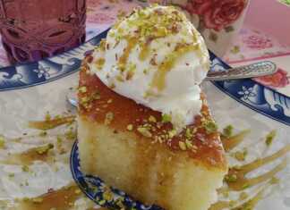 Ένα κομμάτι ραβανί, σιροπιασμένο, με χρυσαφένια επιφάνεια, σε ένα πιάτο, δίπλα σε μια μπάλα παγωτό, περιχυμένο με σιρόπι καραμέλας και πασπαλισμένο με ψιλοκομμένο φιστίκι.