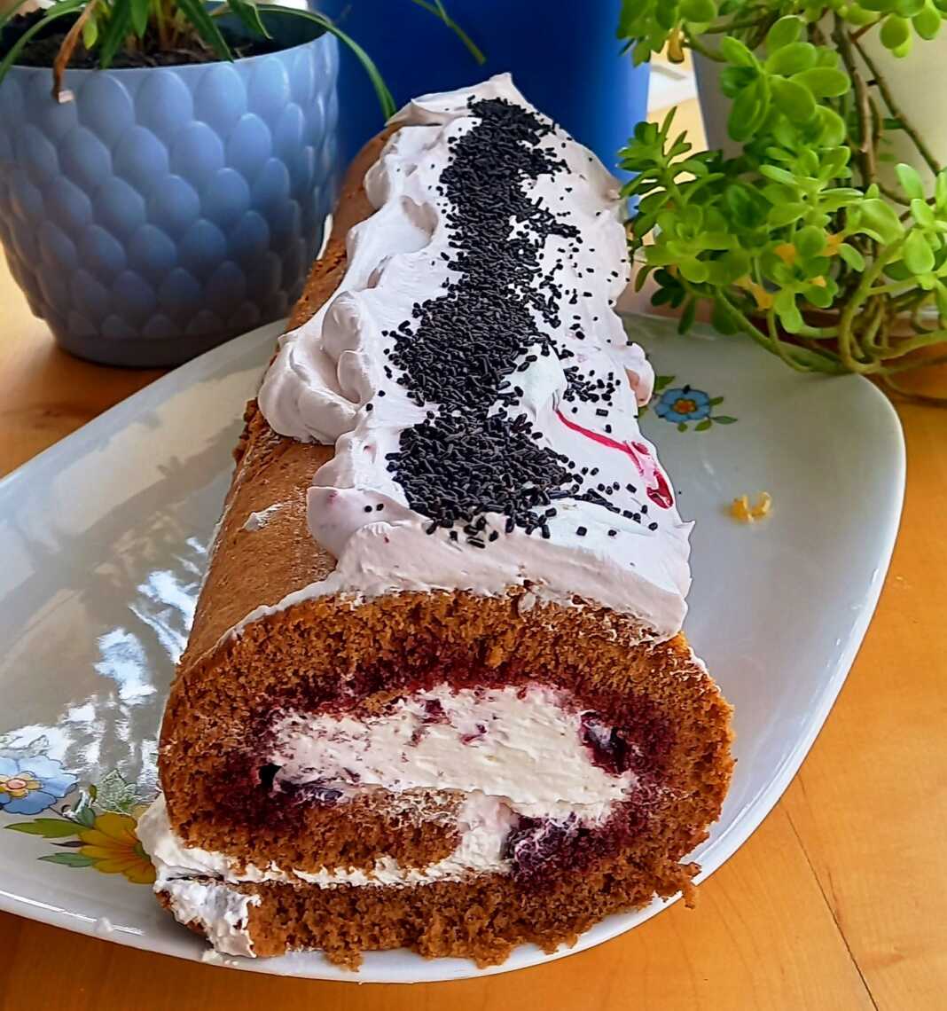 ρολό σοκολατένιο παντεσπάνι γεμιστό με λευκή κρέμα και κόκκινο γλυκό κεράσι, σε ένα πιάτο.