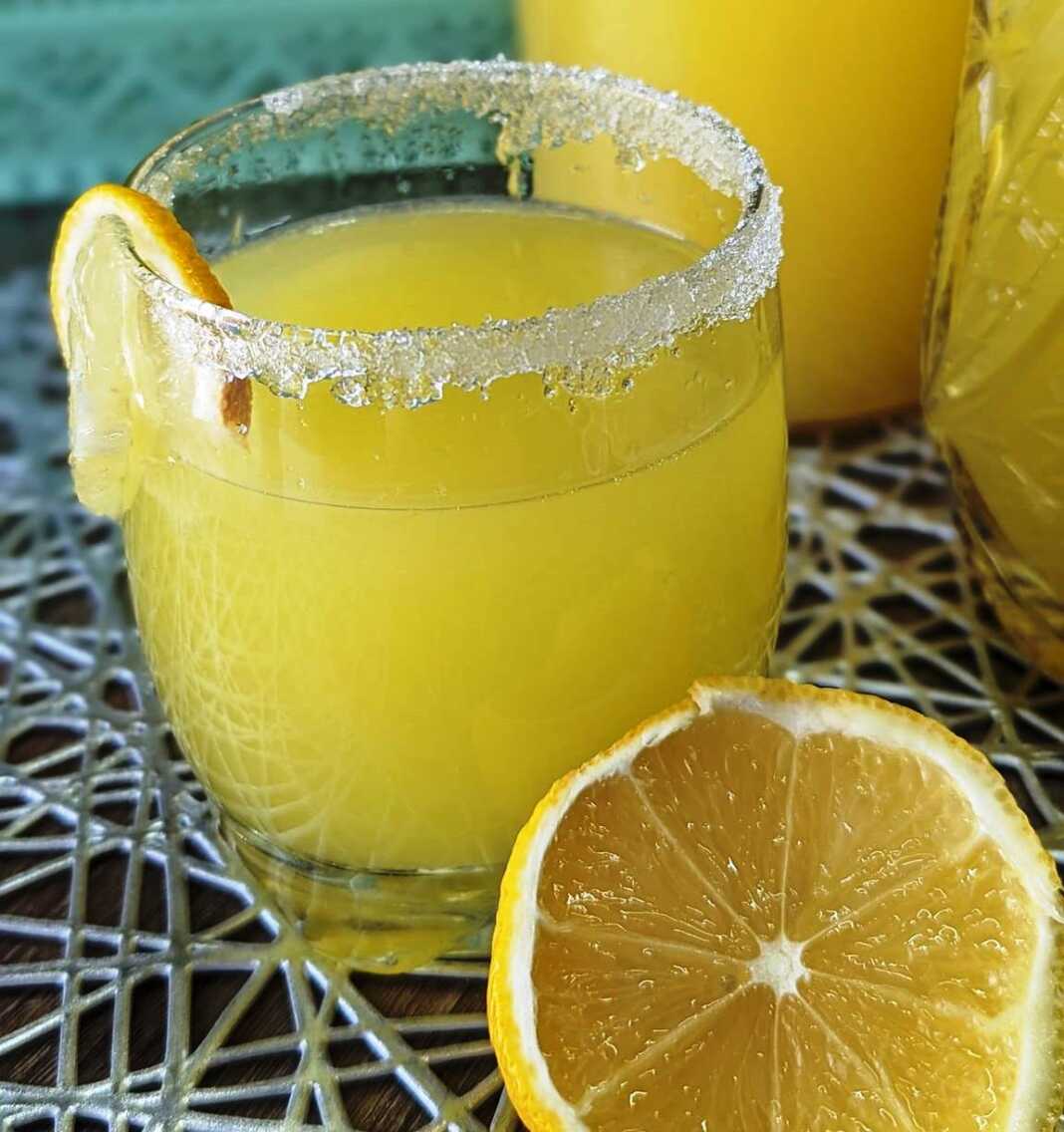 Μια κανάτα γεμάτη κιτρινωπή λεμονάδα με κομματάκια φρούτων και φυσαλίδες.