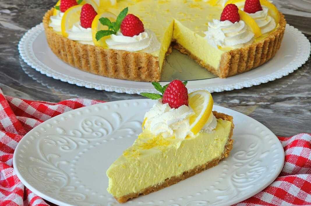 tarta lemoniou Ένα κομμάτι κρεμώδης τάρτα λεμονιού με μπισκοτένια βάση, κρεμώδη κίτρινη γέμιση, γαρνιρισμένη με φρέσκα βατόμουρα και δυόσμο, σερβιρισμένο σε ένα λευκό πιάτο.