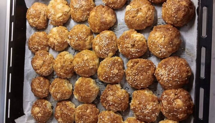 Ένα ταψί γεμάτο με χρυσαφένια, στρογγυλά τυροπιτάκια φούρνου, πασπαλισμένα με σουσάμι, έτοιμα να σερβιριστούν.