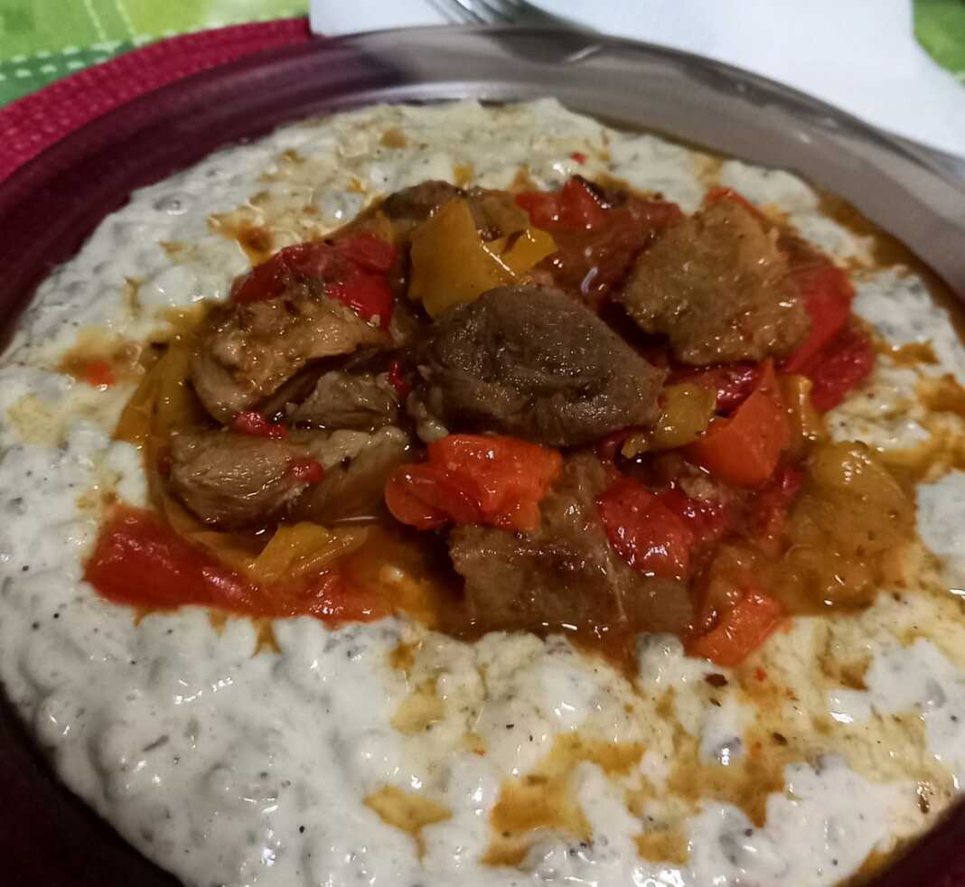 xoirino poures melitzanas0 Ένα πιάτο με κομμάτια χοιρινού ανάμεσα σε χρωματιστές πιπεριές και δίπλα μια μεγάλη κουταλιά από έναν κρεμώδη, γκριζωπό πουρέ.