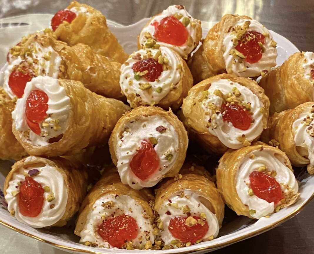 Πιάτο γεμάτο με γλυκά κορνεδάκια, γεμισμένα με λευκή κρέμα και γαρνιρισμένα με κόκκινο γλυκό του κουταλιού και ψιλοκομμένο φιστίκι, έτοιμα για σερβίρισμα.