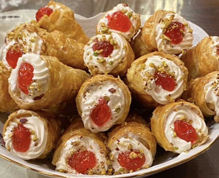 Πιάτο γεμάτο με γλυκά κορνεδάκια, γεμισμένα με λευκή κρέμα και γαρνιρισμένα με κόκκινο γλυκό του κουταλιού και ψιλοκομμένο φιστίκι, έτοιμα για σερβίρισμα.