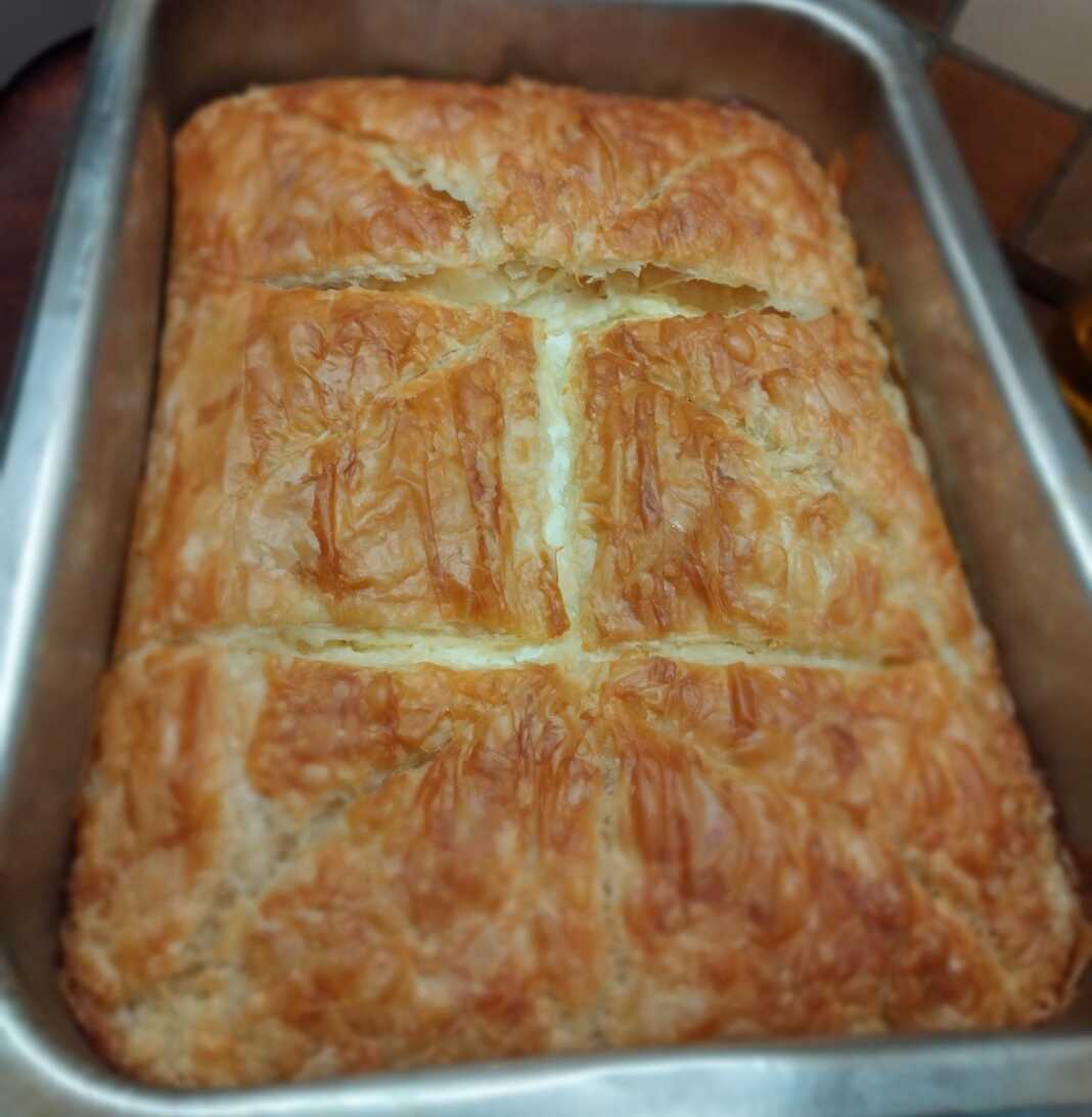 Μια χρυσαφένια τυρόπιτα με μπεσαμέλ, φέτα και κότατζ, κομμένη σε κομμάτια, έτοιμη για σερβίρισμα