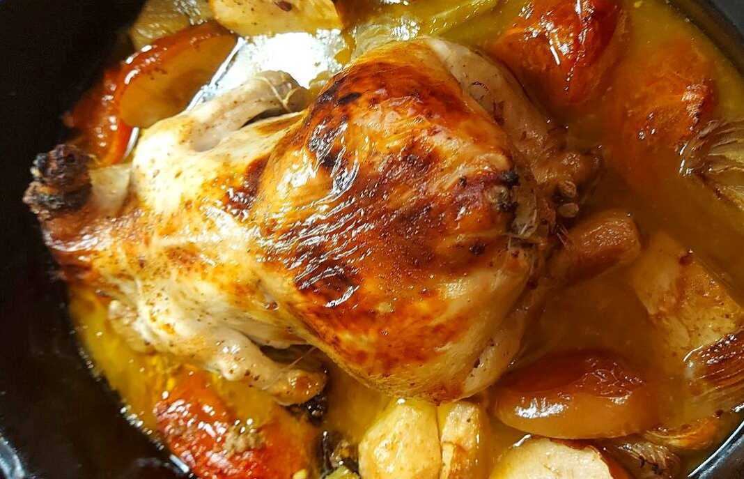 Ένα ολόκληρο ροδοκοκκινισμένο κοτόπουλο γεμιστό μέσα στη γάστρα περιτριγυρισμένο από μήλα, δαμάσκηνα και αρωματικά.