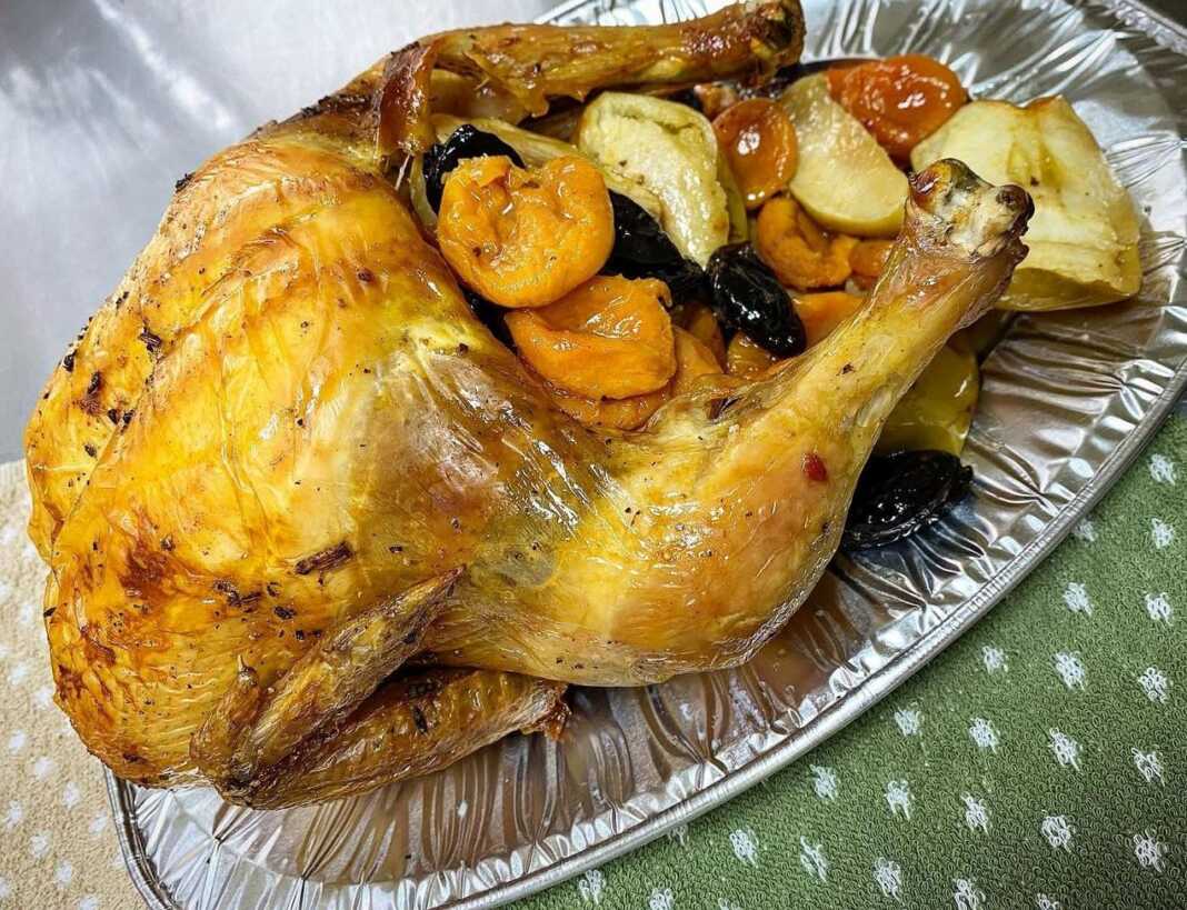 Ολόκληρο ψημένο κοτόπουλο με χρυσαφένια κρούστα, τοποθετημένο σε ταψί με ψημένα κομμάτια μήλων και αποξηραμένα βερίκοκα.