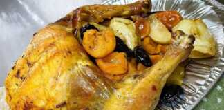 Ολόκληρο ψημένο κοτόπουλο με χρυσαφένια κρούστα, τοποθετημένο σε ταψί με ψημένα κομμάτια μήλων και αποξηραμένα βερίκοκα.