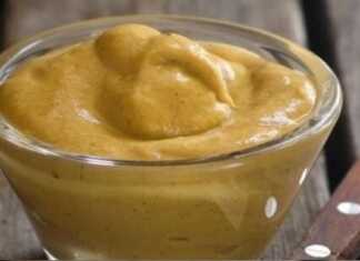 Ένα μικρό γυάλινο βαζάκι γεμάτο με φρέσκια, κίτρινη σπιτική μουστάρδα δίπλα σε λεμόνια και σκόνη μουστάρδας.