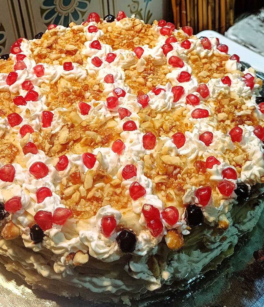 Μια επιβλητική τούρτα με ροζέτες σαντιγής, καλυμμένη με καραμελωμένα αμύγδαλα, σπόρους ροδιού και γλυκό κουταλιού αρώνια πάνω σε γιορτινή πιατέλα.