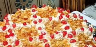 Μια επιβλητική τούρτα με ροζέτες σαντιγής, καλυμμένη με καραμελωμένα αμύγδαλα, σπόρους ροδιού και γλυκό κουταλιού αρώνια πάνω σε γιορτινή πιατέλα.