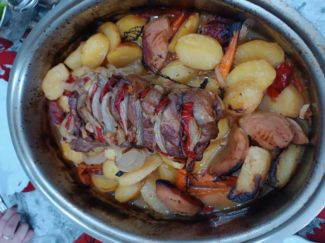 Ένα καλοψημένο χοιρινό ρολό τοποθετημένο πάνω σε μια πανδαισία λαχανικών με πατάτες, καρότα και κυδώνια μέσα σε πήλινη γάστρα.