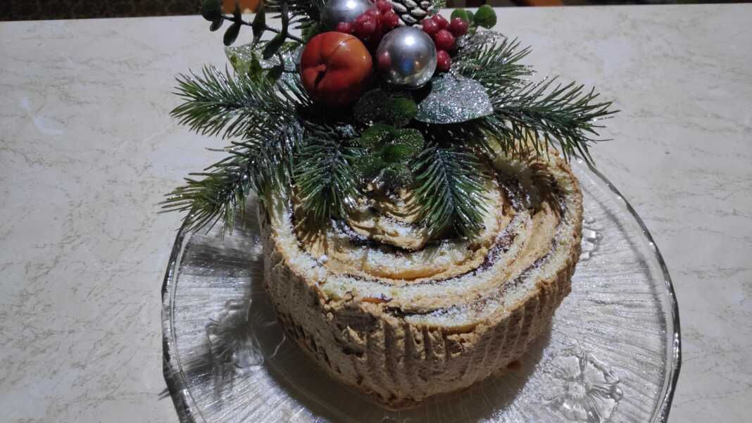 Ένα εντυπωσιακό χριστουγεννιάτικο κέικ κορμός με επικάλυψη σοκολάτας και σχέδια φλοιού δέντρου, στολισμένο γιορτινά.