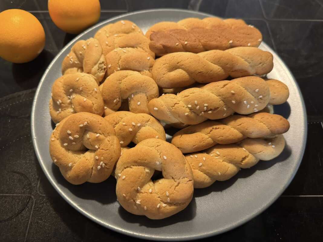 Ένα μπολ γεμάτο με χρυσαφένια, παραδοσιακά κουλουράκια πορτοκαλιού, δίπλα σε ένα φλιτζάνι καφέ και φέτες πορτοκαλιού.