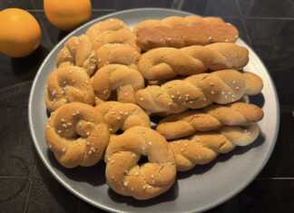 Ένα μπολ γεμάτο με χρυσαφένια, παραδοσιακά κουλουράκια πορτοκαλιού, δίπλα σε ένα φλιτζάνι καφέ και φέτες πορτοκαλιού.