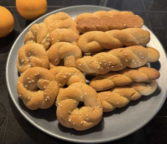 Ένα μπολ γεμάτο με χρυσαφένια, παραδοσιακά κουλουράκια πορτοκαλιού, δίπλα σε ένα φλιτζάνι καφέ και φέτες πορτοκαλιού.