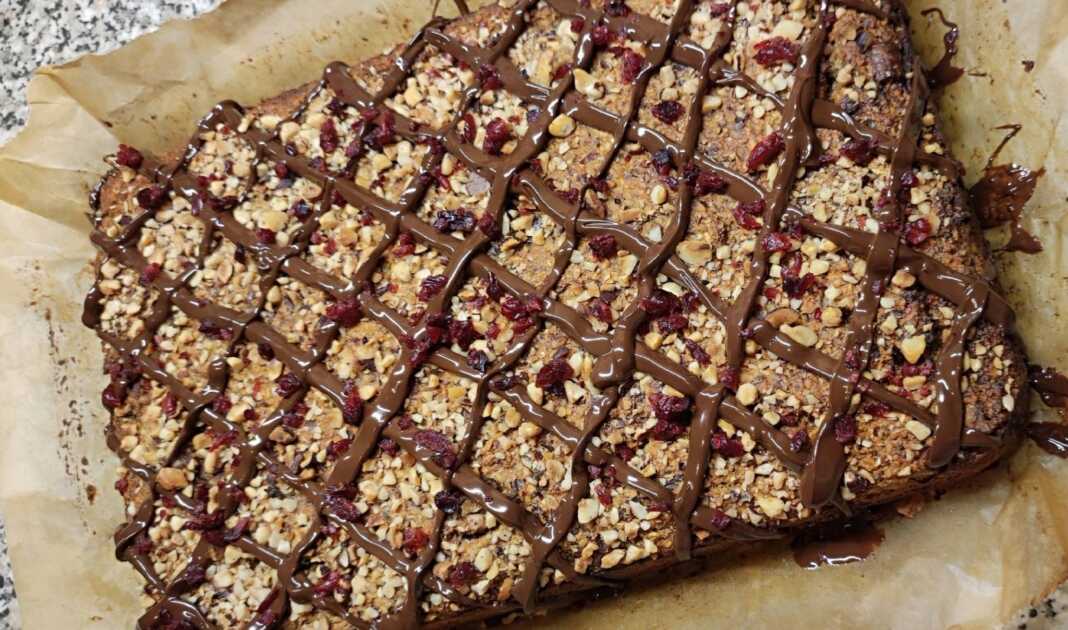 Ένα λαχταριστό κέικ υγείας με επικάλυψη μαύρης σοκολάτας και ξηρούς καρπούς, κομμένο σε φέτες πάνω σε ξύλινη επιφάνεια.