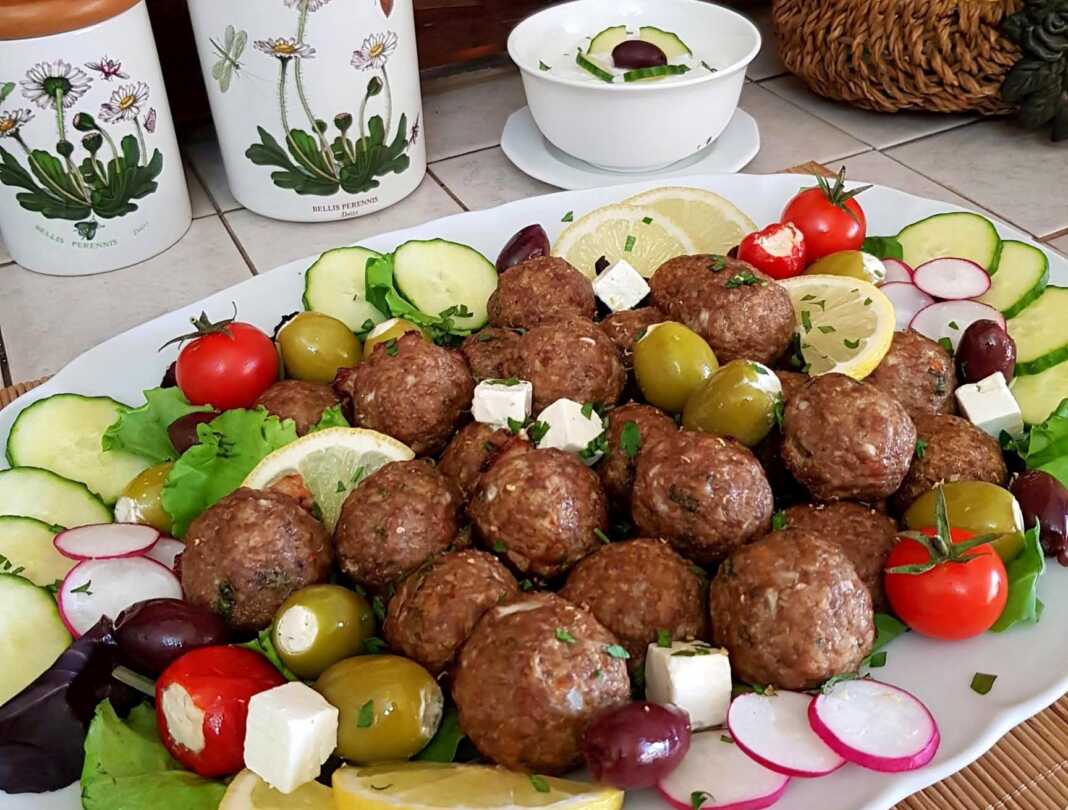 Μια πιατέλα με καλοψημένα αρνίσια κεφτεδάκια, στολισμένα με φρέσκο μαϊντανό, δίπλα σε ένα μπολ με κρεμώδες τζατζίκι και φέτες λεμονιού.