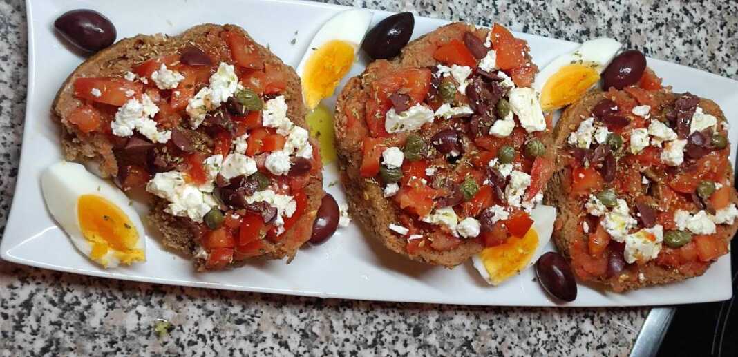 Ένας πλούσιος κρητικός ντάκος πάνω σε ξύλινη επιφάνεια, φορτωμένος με τριμμένη ντομάτα, φέτα, ελιές και ένα βραστό αυγό κομμένο στη μέση.