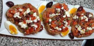 Ένας πλούσιος κρητικός ντάκος πάνω σε ξύλινη επιφάνεια, φορτωμένος με τριμμένη ντομάτα, φέτα, ελιές και ένα βραστό αυγό κομμένο στη μέση.