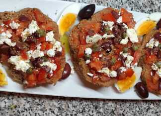 Ένας πλούσιος κρητικός ντάκος πάνω σε ξύλινη επιφάνεια, φορτωμένος με τριμμένη ντομάτα, φέτα, ελιές και ένα βραστό αυγό κομμένο στη μέση.