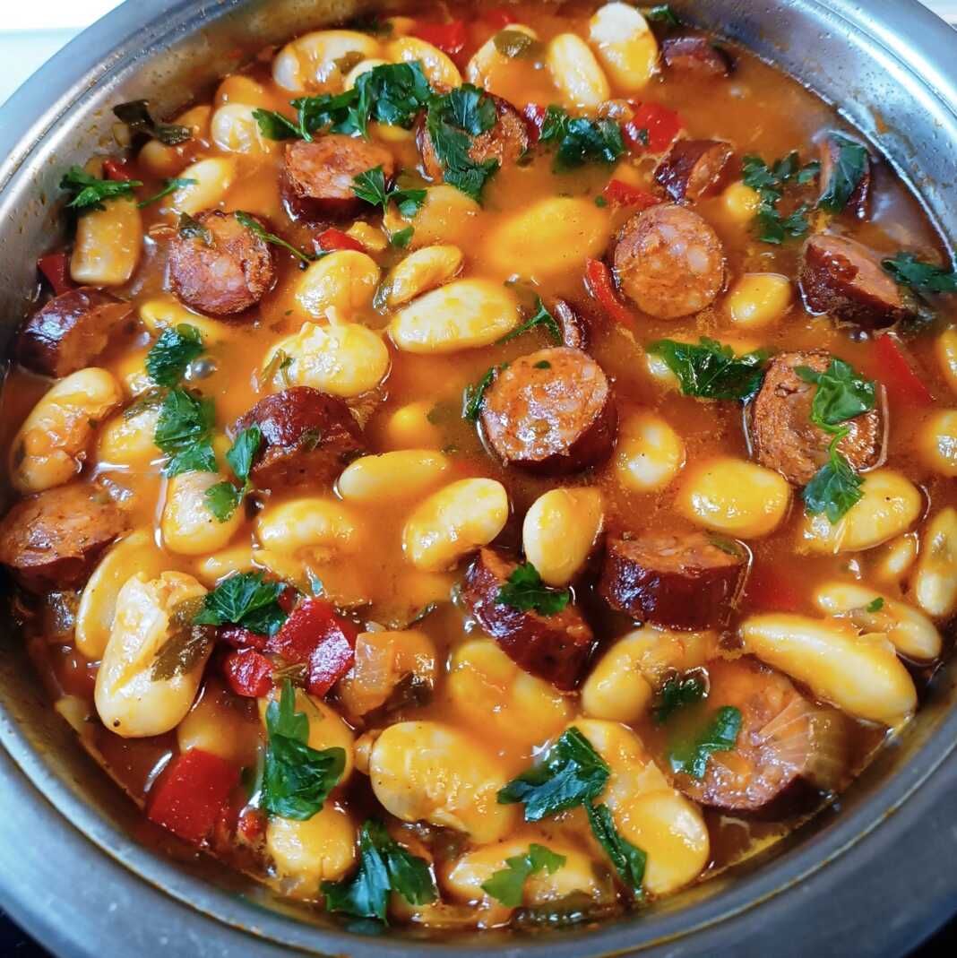 Μια βαθιά κατσαρόλα με μελωμένους γίγαντες σε κόκκινη σάλτσα, με ροδέλες από λουκάνικο και ψιλοκομμένο μαϊντανό στην κορυφή.