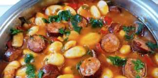 Γίγαντες στην Κατσαρόλα με Λουκάνικο: Το Έξυπνο και Μελωμένο Γεύμα της Εβδομάδας! Μια βαθιά κατσαρόλα με μελωμένους γίγαντες σε κόκκινη σάλτσα, με ροδέλες από λουκάνικο και ψιλοκομμένο μαϊντανό στην κορυφή.