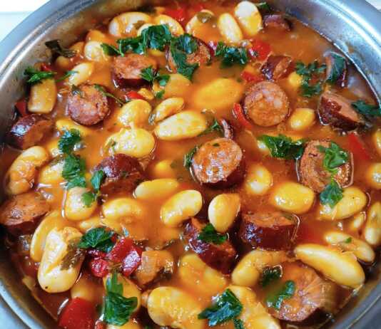 Μια βαθιά κατσαρόλα με μελωμένους γίγαντες σε κόκκινη σάλτσα, με ροδέλες από λουκάνικο και ψιλοκομμένο μαϊντανό στην κορυφή.