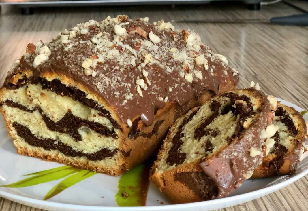 Ένα μακρόστενο μαρμάρινο κέικ με όμορφα νερά σοκολάτας και βανίλιας, καλυμμένο με γυαλιστερό γλάσο σοκολάτας και τριμμένους ξηρούς καρπούς.