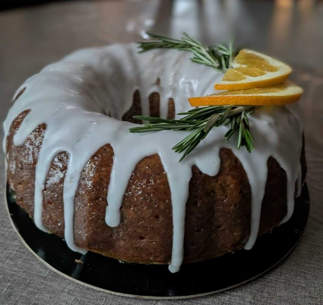 Ένα ολόκληρο σιροπιασμένο κέικ αμυγδάλου πασπαλισμένο με ζάχαρη άχνη, τοποθετημένο σε μια σχάρα, με φέτες πορτοκαλιού δίπλα.