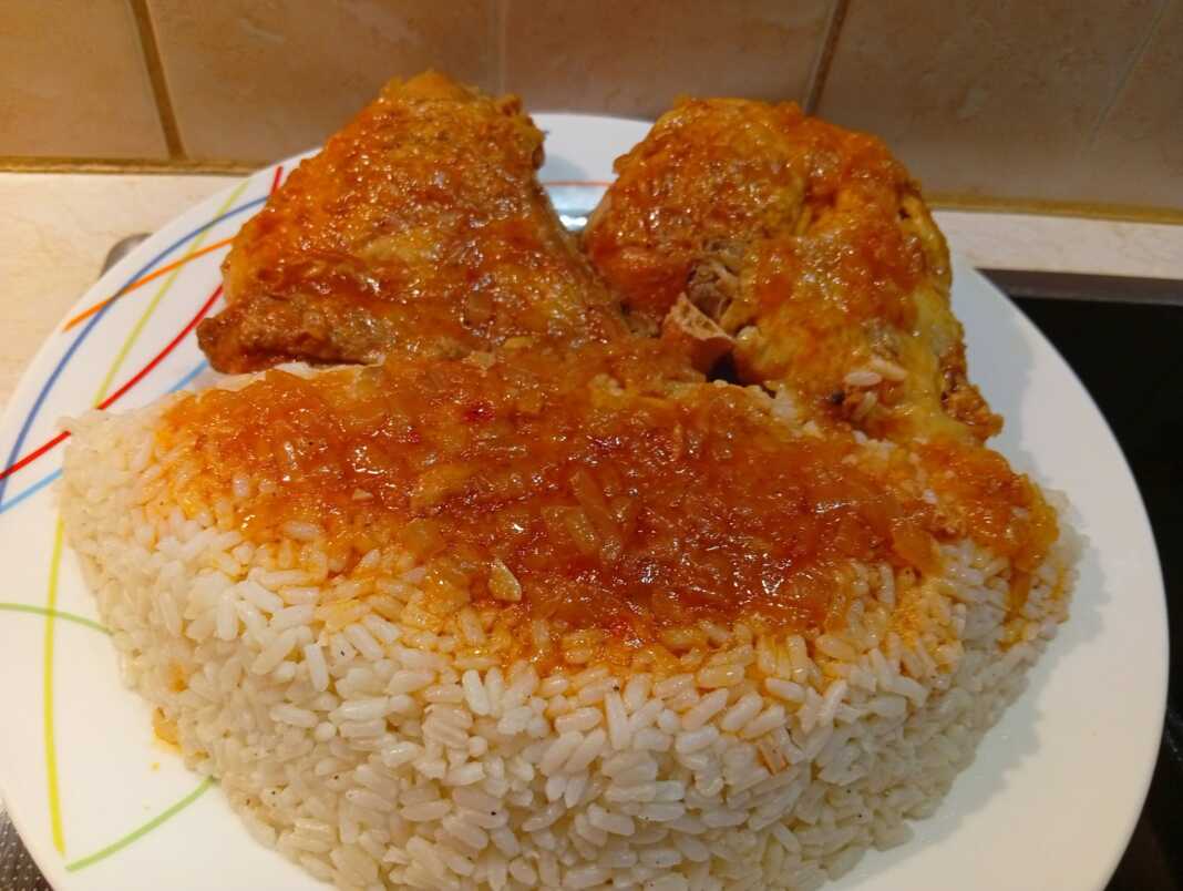 Ένα πιάτο με κομμάτια κοκκινιστού κόκορα σε πλούσια κόκκινη σάλτσα, σερβιρισμένο δίπλα σε ένα λευκό σπυρωτό πιλάφι.