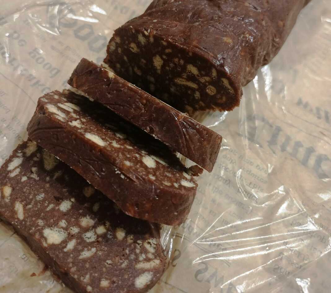 Ένας λαχταριστός σοκολατένιος κορμός τυλιγμένος σε μεμβράνη και ένας κομμένος σε φέτες, δείχνοντας τα κομμάτια μπισκότου στο εσωτερικό του.