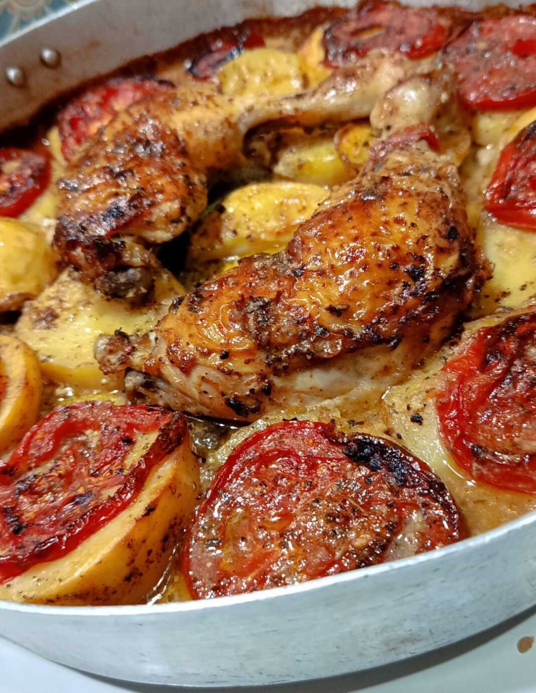 Ένα ταψί με ροδοκοκκινισμένα μπούτια κοτόπουλο, πατάτες κομμένες σε ροδέλες και ψημένες φέτες ντομάτας, περιχυμένα με σάλτσα μουστάρδας.