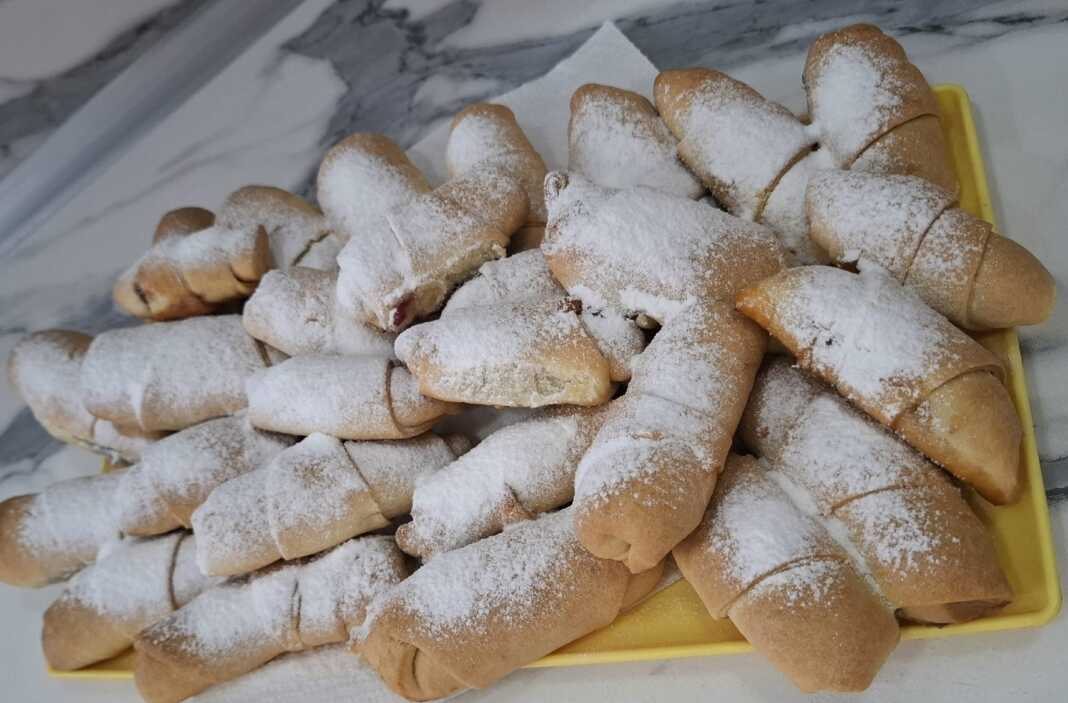 Ένας δίσκος με χρυσαφένια κρουασανάκια πασπαλισμένα με άχνη ζάχαρη, δίπλα σε ένα μπολάκι με μαρμελάδα μήλο.