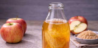 Ένα γυάλινο βάζο με θολό μηλόξυδο όπου στο εσωτερικό του διακρίνεται η ζελατινώδης "μάνα" και δίπλα υπάρχουν φρέσκα κόκκινα μήλα.