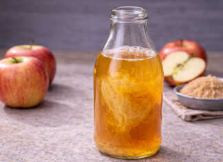 Ένα γυάλινο βάζο με θολό μηλόξυδο όπου στο εσωτερικό του διακρίνεται η ζελατινώδης "μάνα" και δίπλα υπάρχουν φρέσκα κόκκινα μήλα.