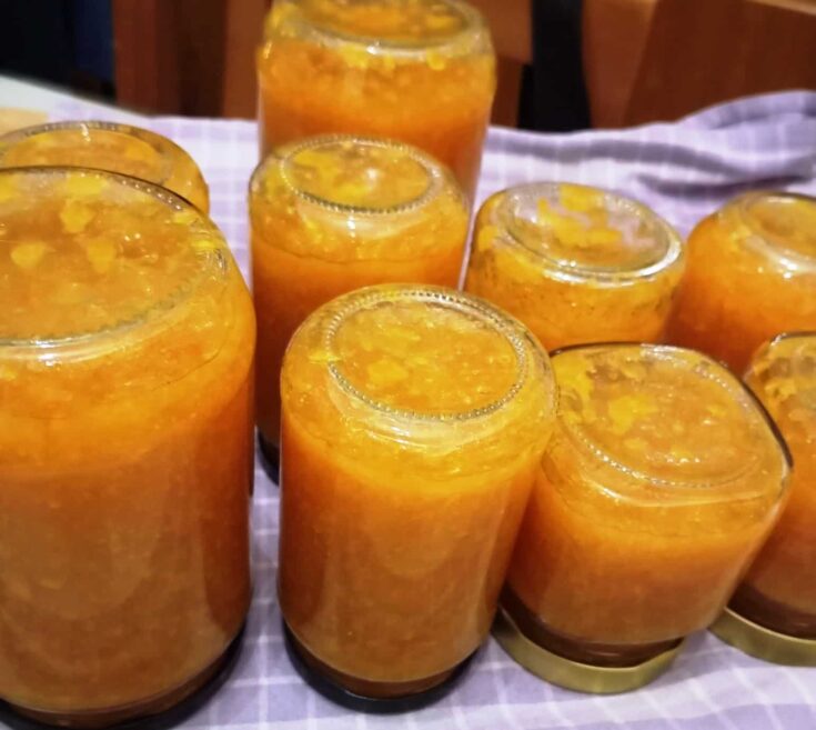 Βάζα γεμάτα με κατακίτρινη, λαμπερή μαρμελάδα πορτοκάλι δίπλα σε φέτες φρέσκων φρούτων πάνω σε έναν ξύλινο πάγκο.